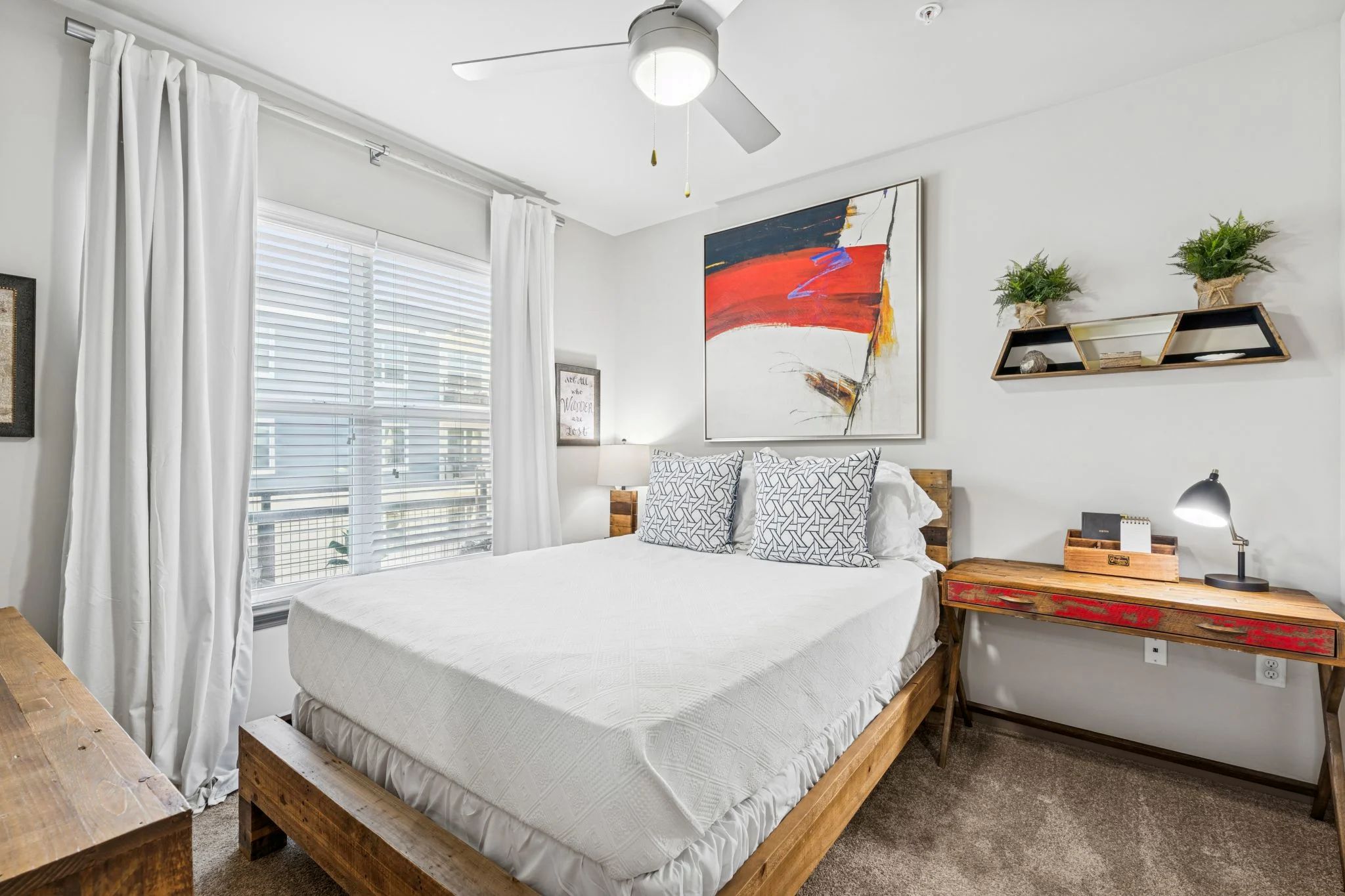 A cozy apartment bedroom featuring a wooden bed frame, white bedding, colorful artwork, and a rustic desk.